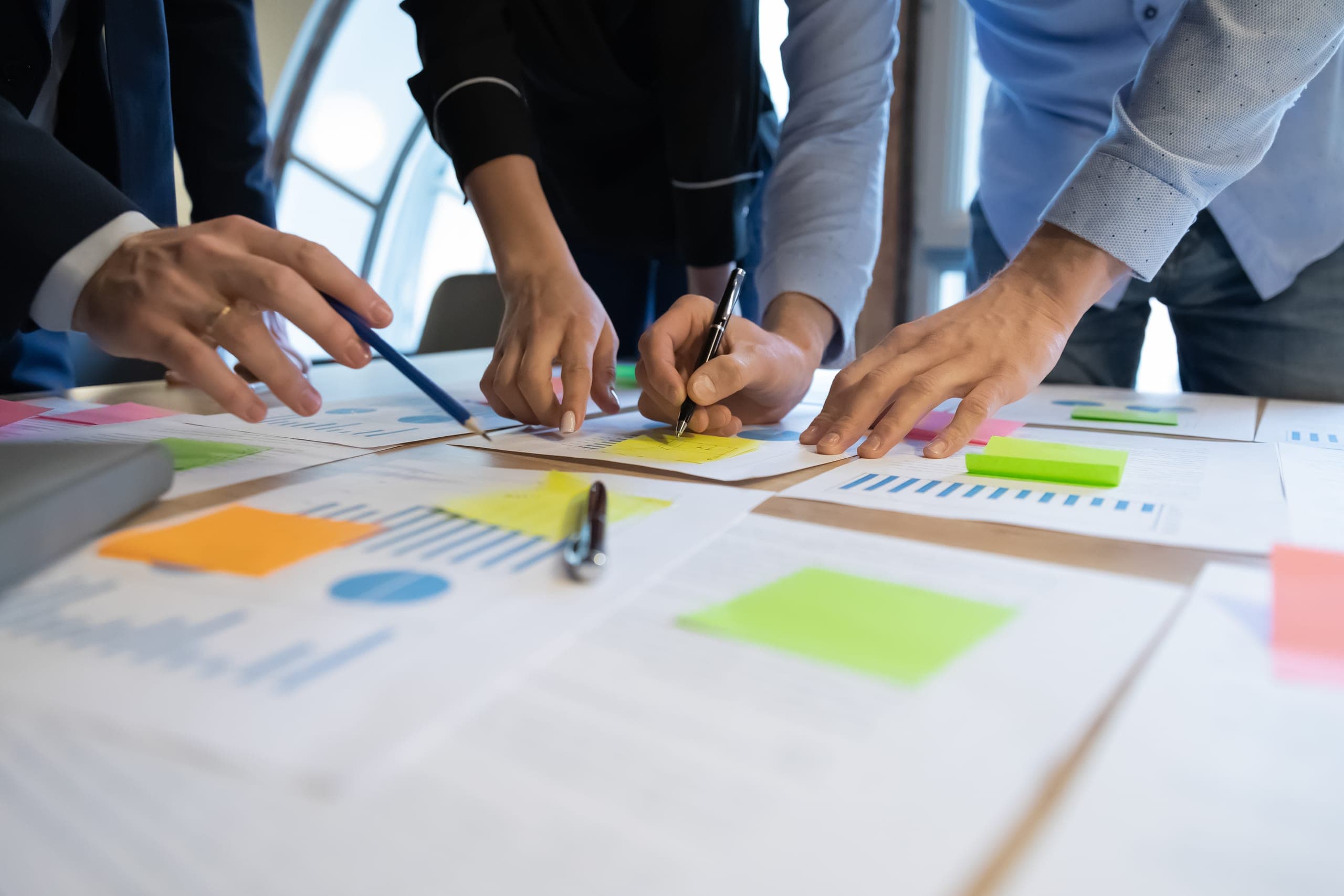 Several people standing around a table, pointing at and annotating printed charts covered in coloured sticky notes